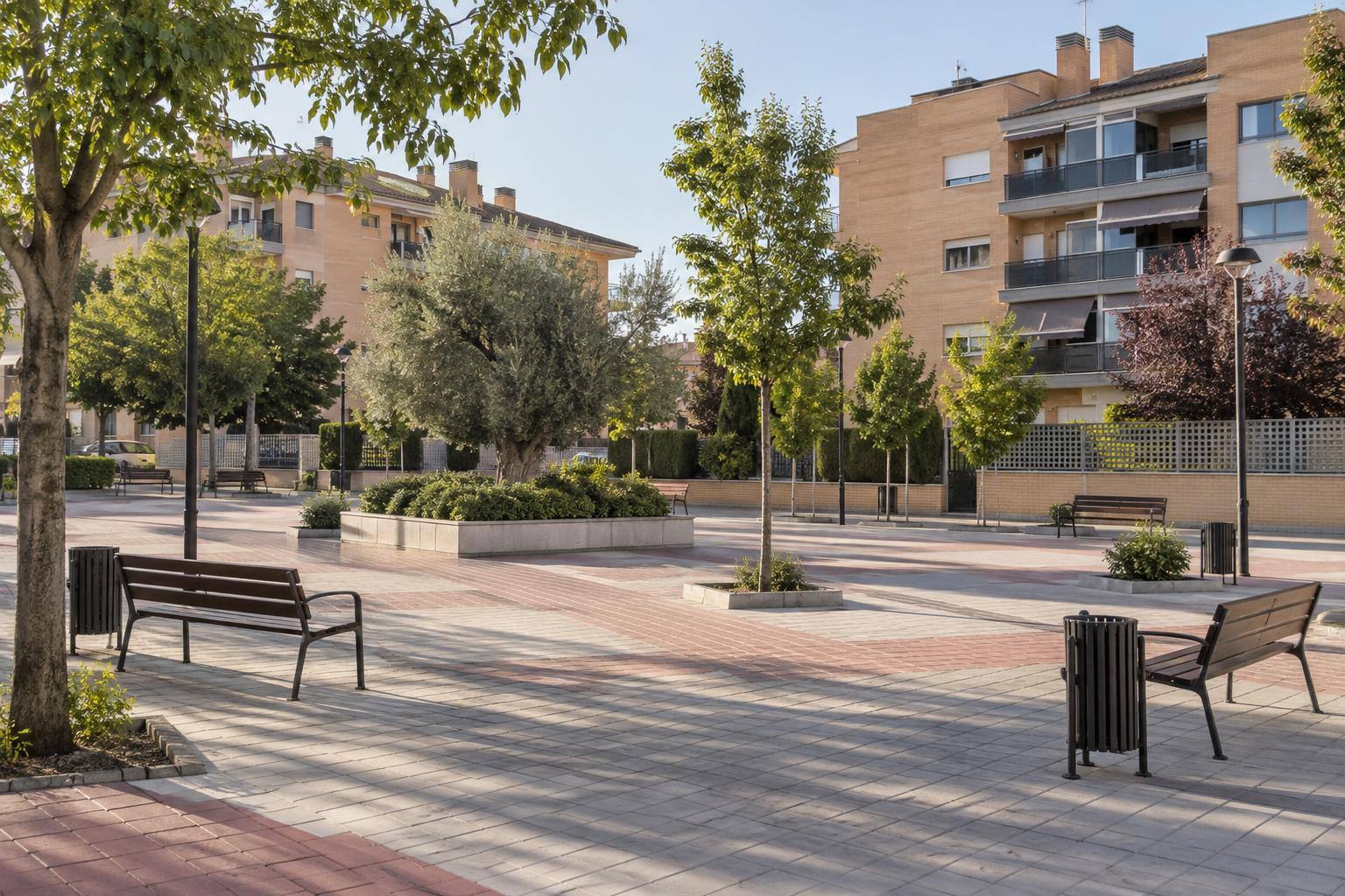Imagen ilustrativa: Plaza de una barriada residencial con bancos, papeleras y aceras, sin personas reconocibles.