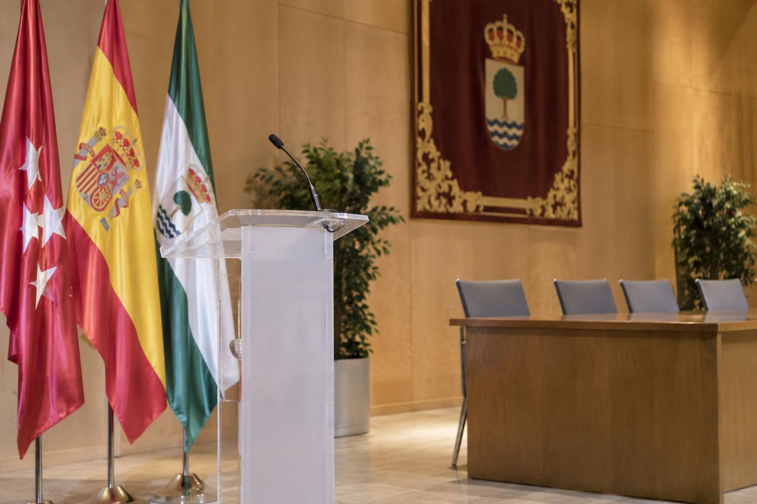 Imagen ilustrativa: Atril con micrófono y sillas en una sala de actos municipal, sin personas ni textos identificables