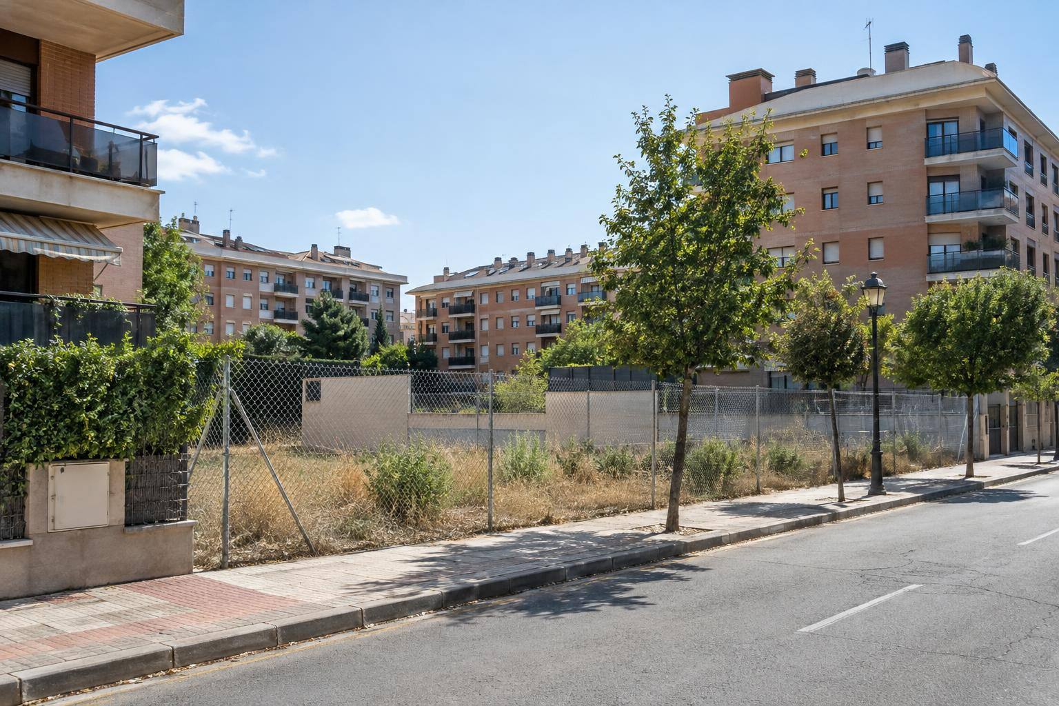 Imagen ilustrativa: Calle de un barrio residencial con edificios de viviendas y una parcela urbana vallada, sin personas reconocibles.