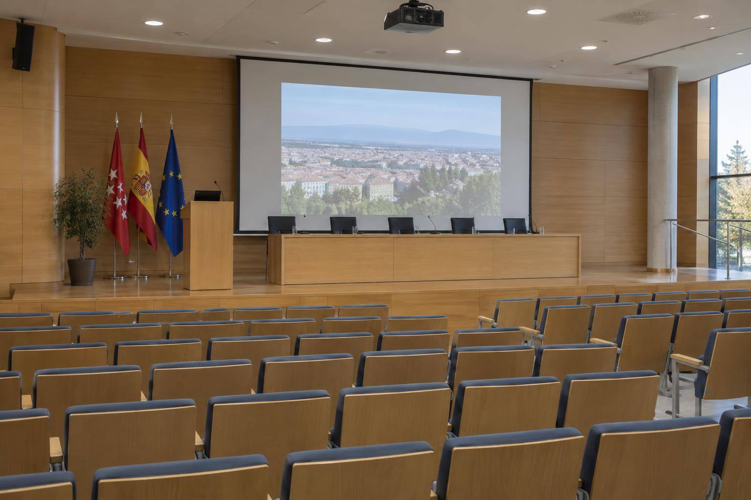 Imagen ilustrativa: Salón de actos universitario con una pantalla de proyección, un atril y filas de butacas vacías.