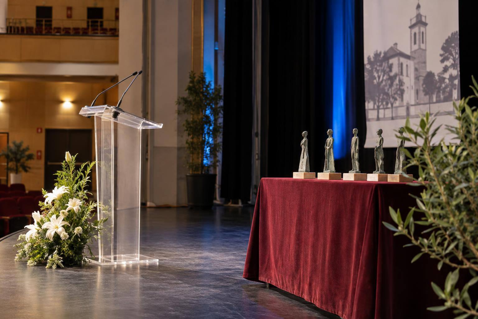 Imagen ilustrativa: Escenario de un teatro con un atril y pequeñas esculturas sobre una mesa para una entrega de premios culturales, sin personas reconocibles.