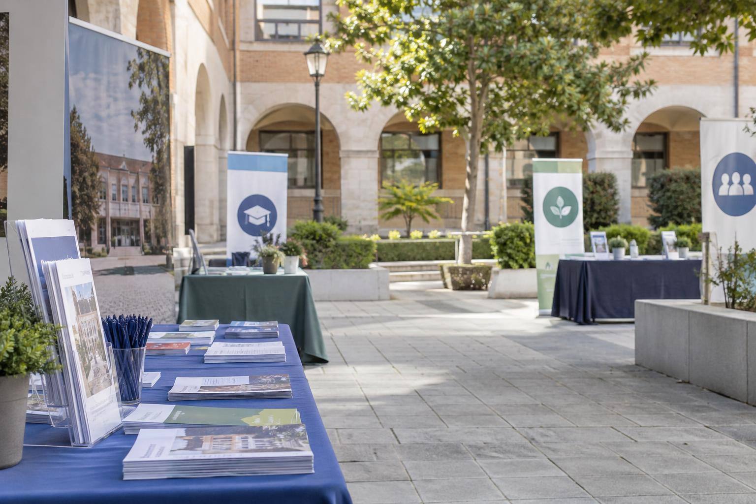 Imagen ilustrativa: Patio universitario con mesas informativas, folletos y expositores de servicios al alumnado, sin personas reconocibles.