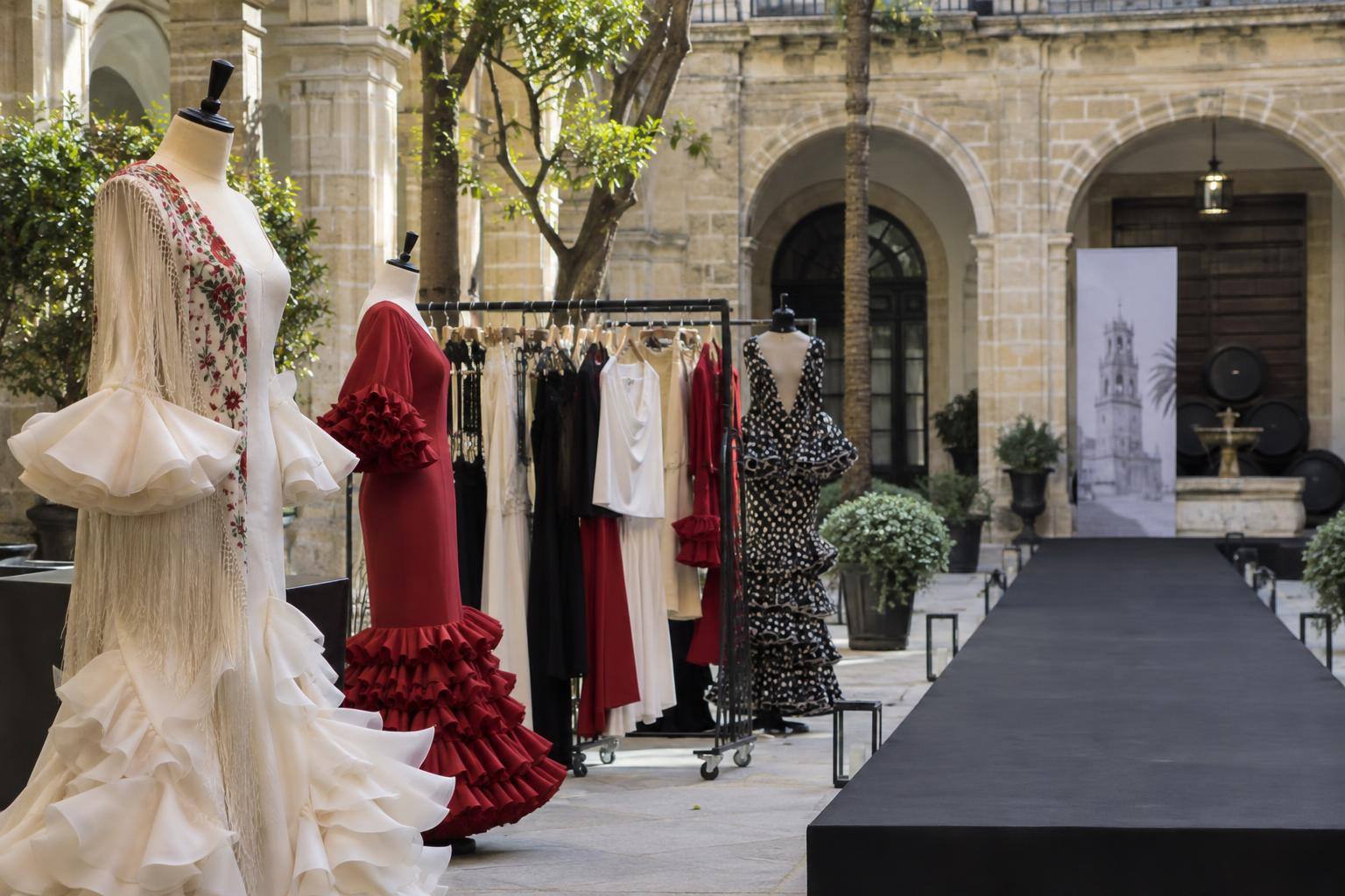 Imagen ilustrativa: Prendas de diseño en percheros y maniquíes junto a una pasarela en un patio histórico, sin personas reconocibles.