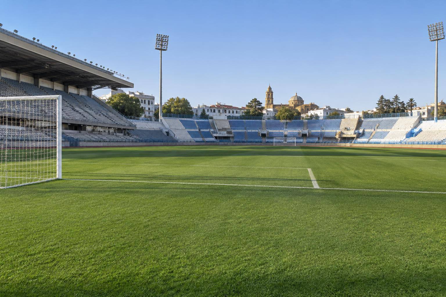 Imagen ilustrativa: Césped de un estadio municipal de fútbol con porterías y gradas vacías, sin personas reconocibles.
