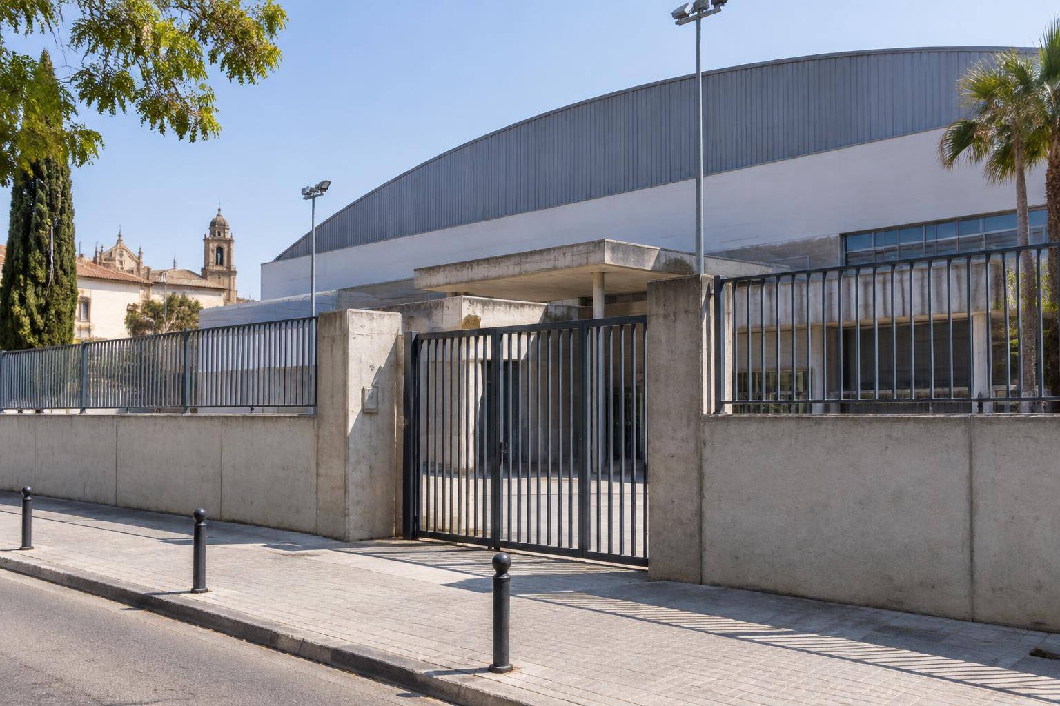 Imagen ilustrativa: Cerramiento de un polideportivo municipal con muro de hormigón, rejas metálicas y una puerta de acceso, sin personas reconocibles.