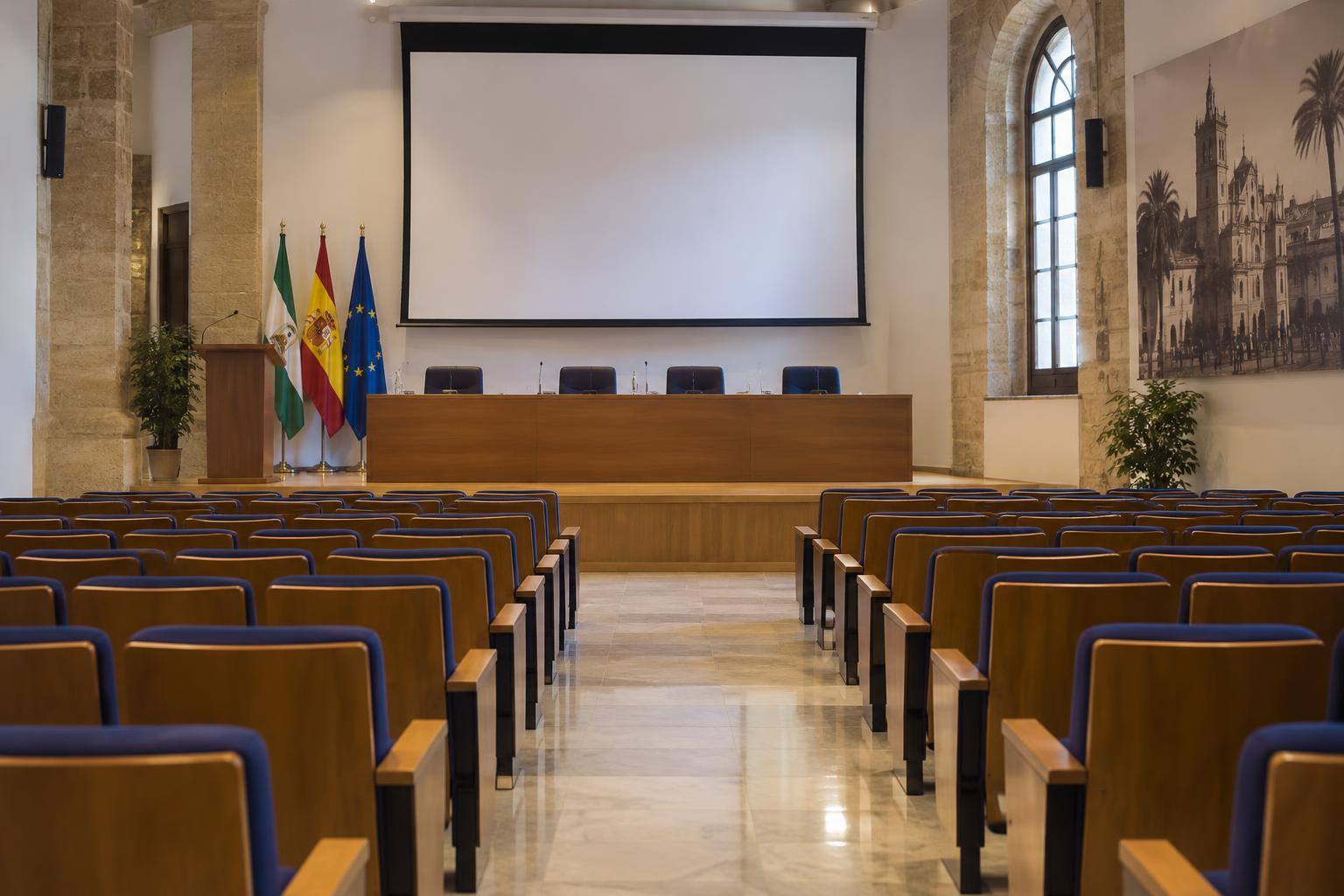 Imagen ilustrativa: Salón de actos universitario vacío con pantalla de proyección y una mesa para conferencias.